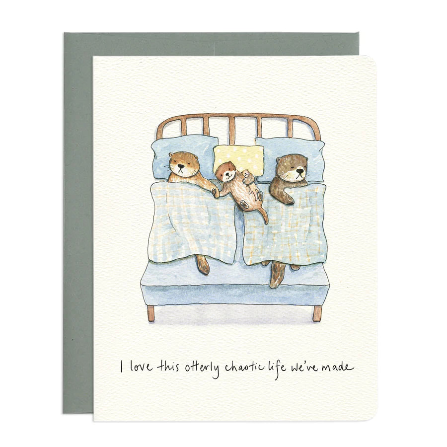 The image features a watercolor illustration of a family of otters in bed. The card itself is white with a textured surface. Under the card is a grey envelope.
The central scene depicts a bed with a light blue mattress and a wooden headboard with simple horizontal slats. Two adult otters and one smaller otter are nestled under light blue blankets with a subtle checkered pattern.
Below the illustration, handwritten in black ink, is the text "I love this otterly chaotic life we've made."