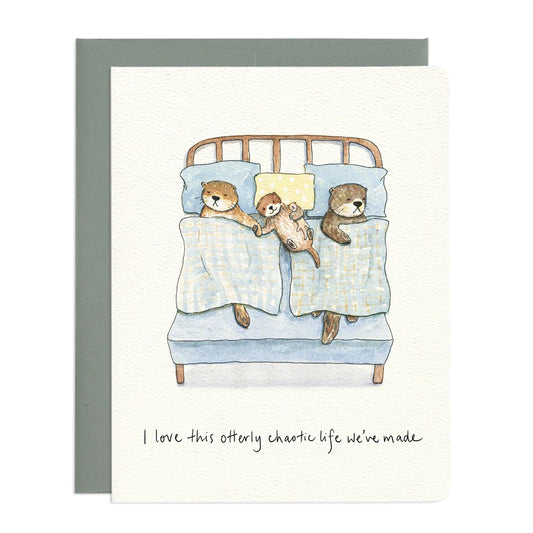 The image features a watercolor illustration of a family of otters in bed. The card itself is white with a textured surface. Under the card is a grey envelope.

The central scene depicts a bed with a light blue mattress and a wooden headboard with simple horizontal slats. Two adult otters and one smaller otter are nestled under light blue blankets with a subtle checkered pattern. 
Below the illustration, handwritten in black ink, is the text "I love this otterly chaotic life we've made." 